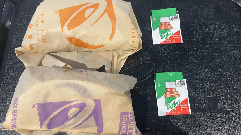 Two wrapped tacos sit on a black tray with Tajín seasoning packets on the side.
