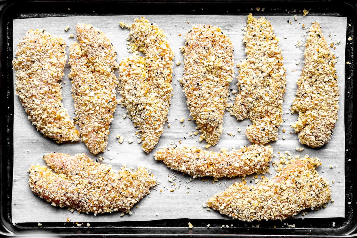 Breaded everything bagel chicken tenders arranged in a single layer on a parchment-lined sheet pan before baking.