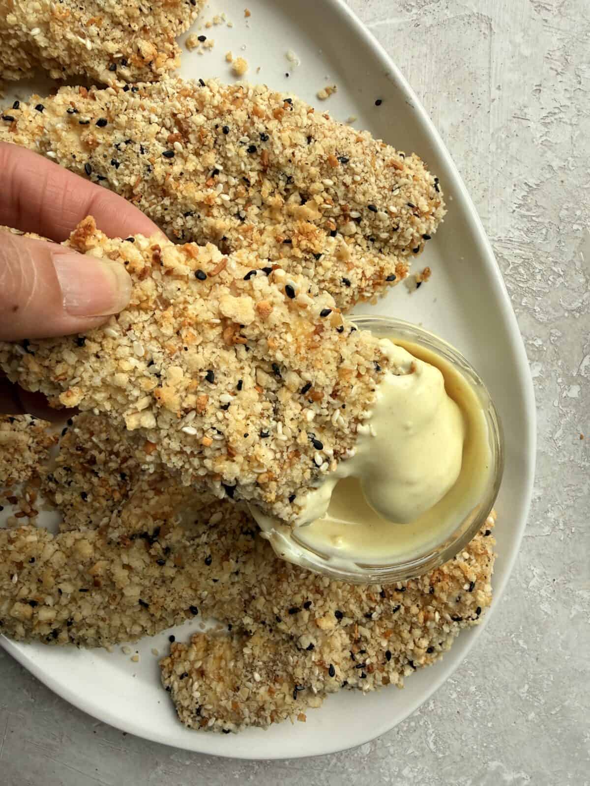 Close-up of an everything bagel chicken tender dipped into sauce, with more tenders piled on the platter behind it.