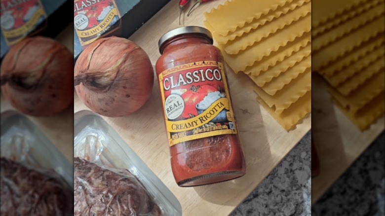 A jar of Classico Creamy Ricotta Pasta Sauce on a counter next to some dry lasagna noodles and an onion