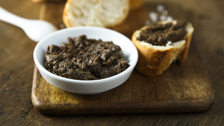 A bowl of olive tapenade next to a piece of baguette topped with it