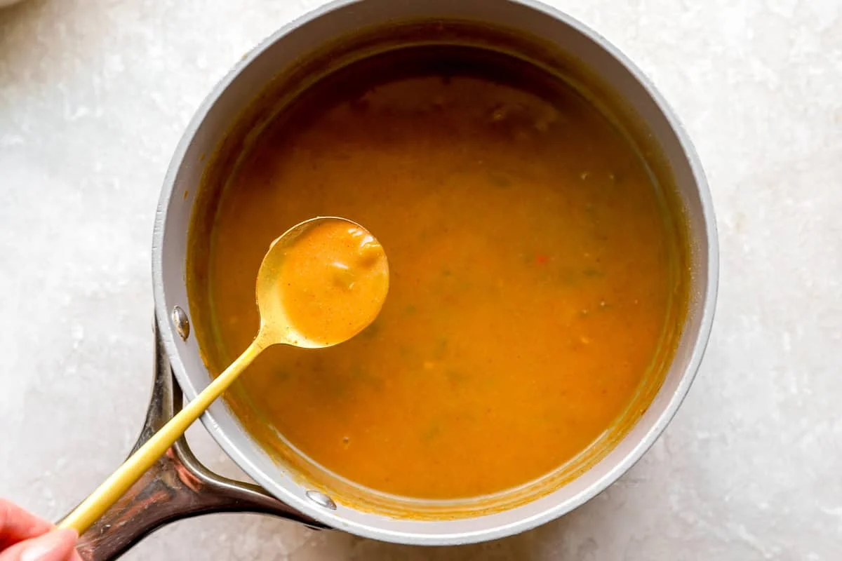 A spoon lifting a scoop of smooth green chile gravy from the saucepan to show its thickened, pourable consistency.