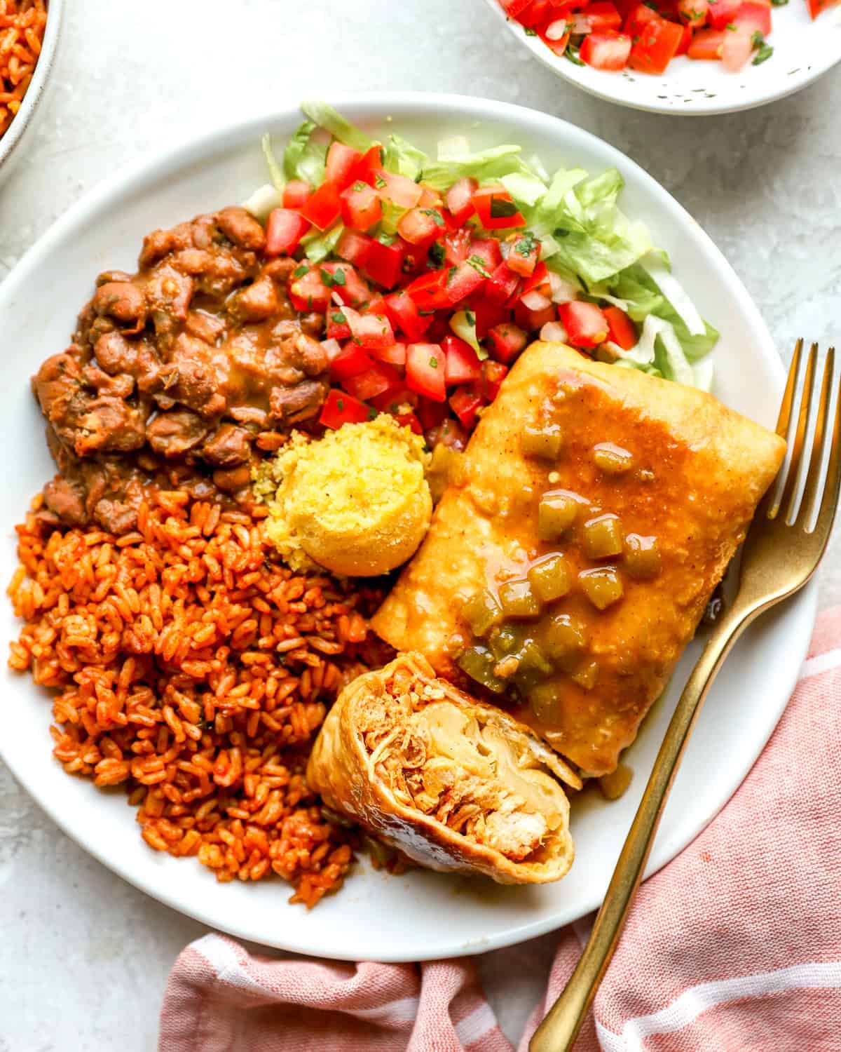 Overhead shot of a Chi-Chi's chimichanga cut in half on a white plate, showing shredded chicken and melted cheese inside, topped with green chile gravy and served with Mexican rice, pinto beans, cornbread, pico de gallo, and shredded lettuce.