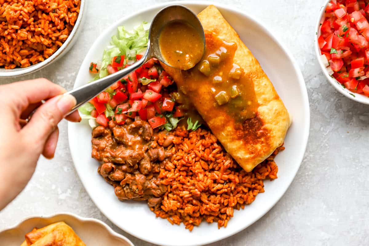 A spoon pours warm green chile gravy over a fried chimichanga plated with Mexican rice, pinto beans, pico de gallo, and shredded lettuce.