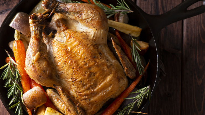Whole roast chicken in a cast iron pan surrounded by vegetables and herbs