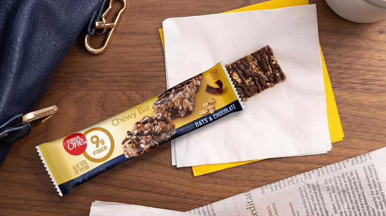 Close-up of a Fiber One Chewy Bar partially unwrapped on a napkin on a desk next to a bag and a newspaper