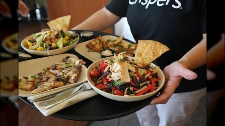 A Crispers employee is holding a tray with various flatbreads and salads.