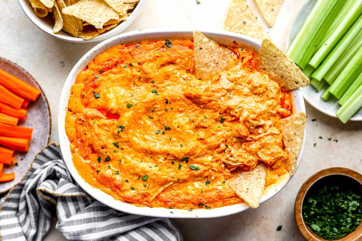Overhead shot of buffalo chicken dip surrounded by tortilla chips, celery sticks, carrot sticks, and chopped herbs.