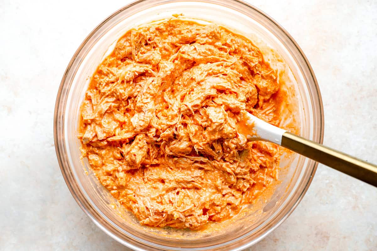 Buffalo chicken dip mixture being stirred together in a glass bowl before baking, streaky and bright orange.