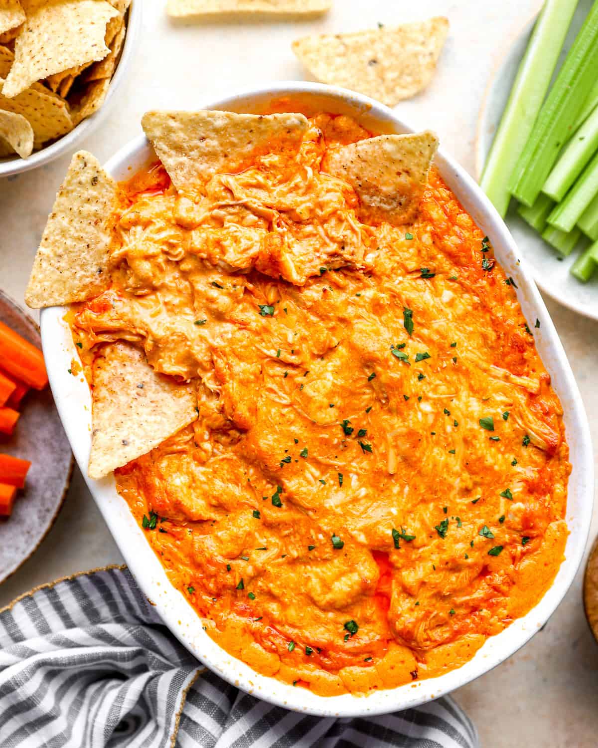 Baked buffalo chicken dip in a white oval baking dish, topped with tortilla chips and fresh herbs, with more chips, celery, and carrots alongside.