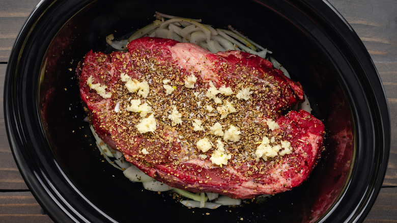 A raw chuck roast with seasonings in a slow cooker