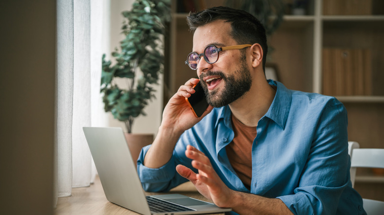 Bearded man with glasses working from home on laptop while talking on his mobile phone