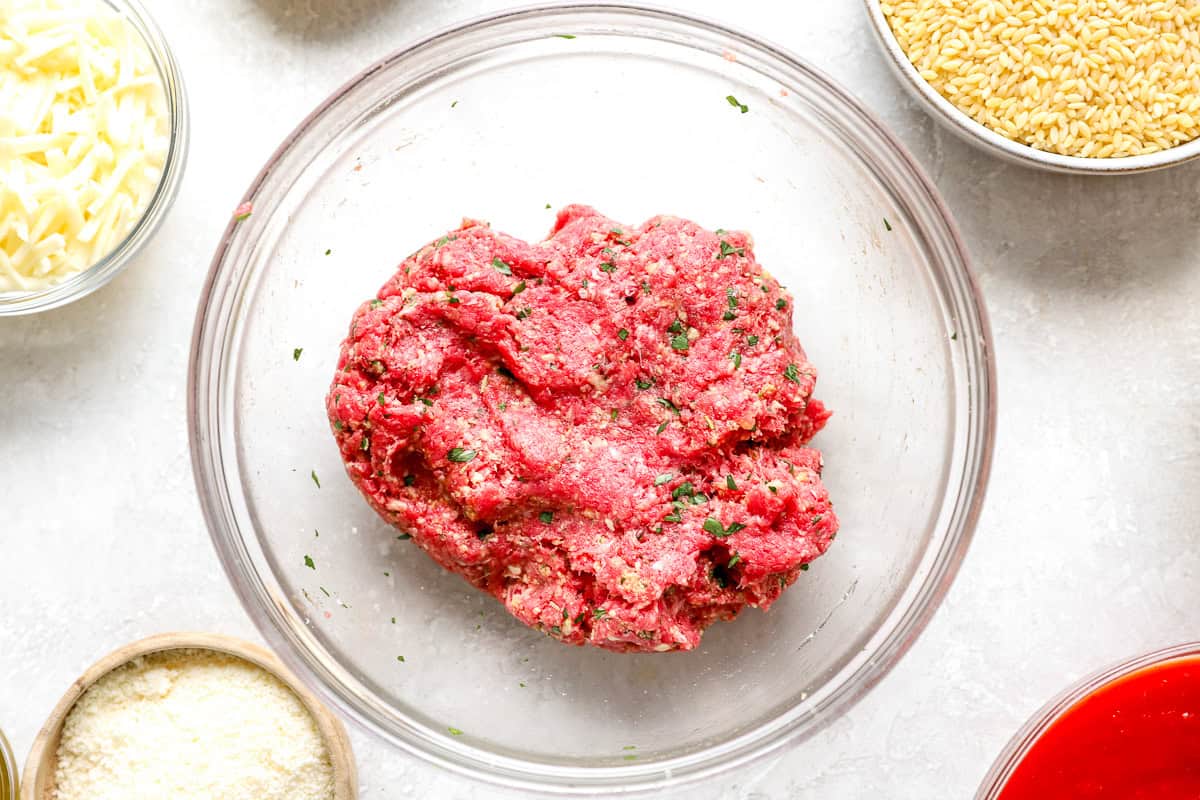 Raw ground beef meatball mixture in a glass bowl, surrounded by shredded mozzarella, dry orzo, and marinara sauce.