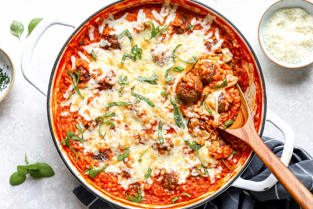 Baked meatball parmesan orzo with golden bubbly cheese and fresh basil, a wooden spoon scooping a serving.