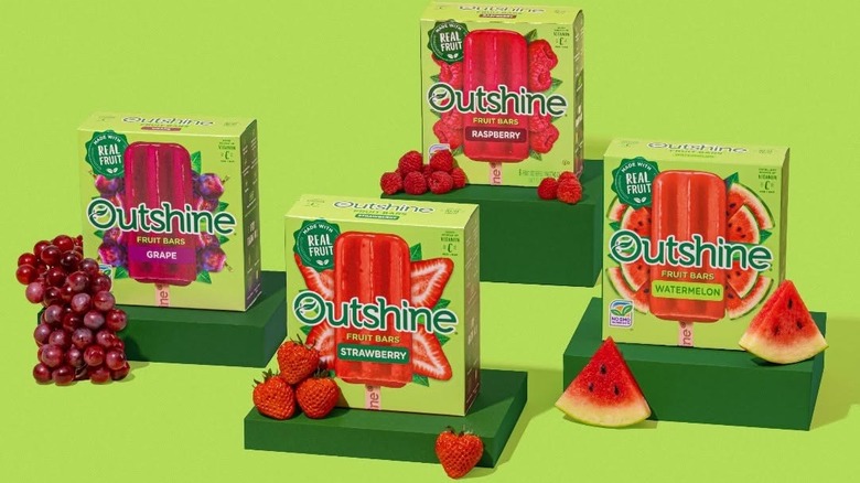 Four boxes of Outshine Fruit Bars on green pedestals with various fruits against a green background