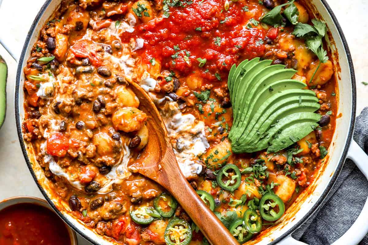Close-up of taco gnocchi skillet with melted cheese, sour cream, salsa, sliced avocado, and jalapeños.