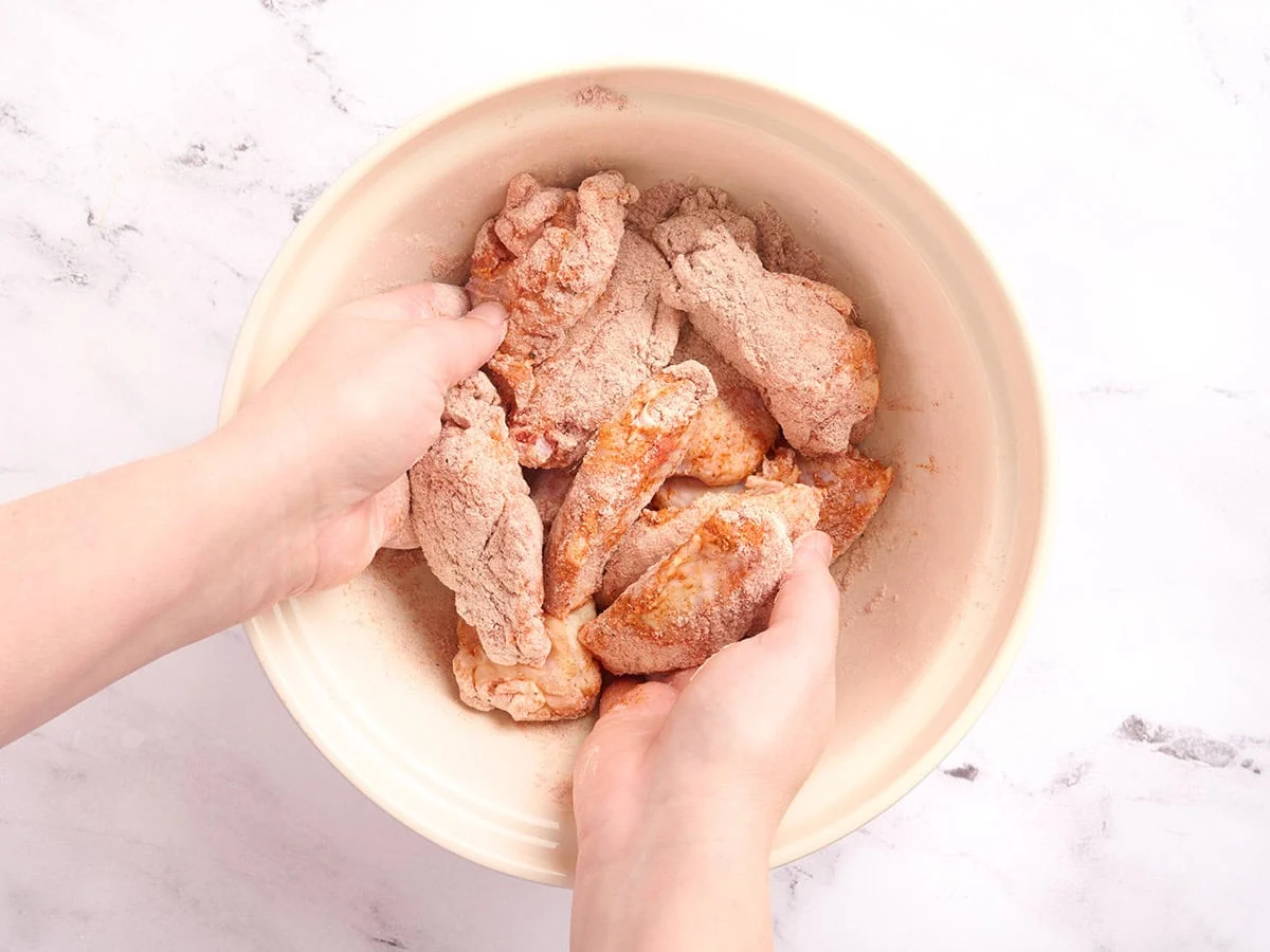 Hands tossing raw chicken wings in the seasoned flour mixture until fully coated.