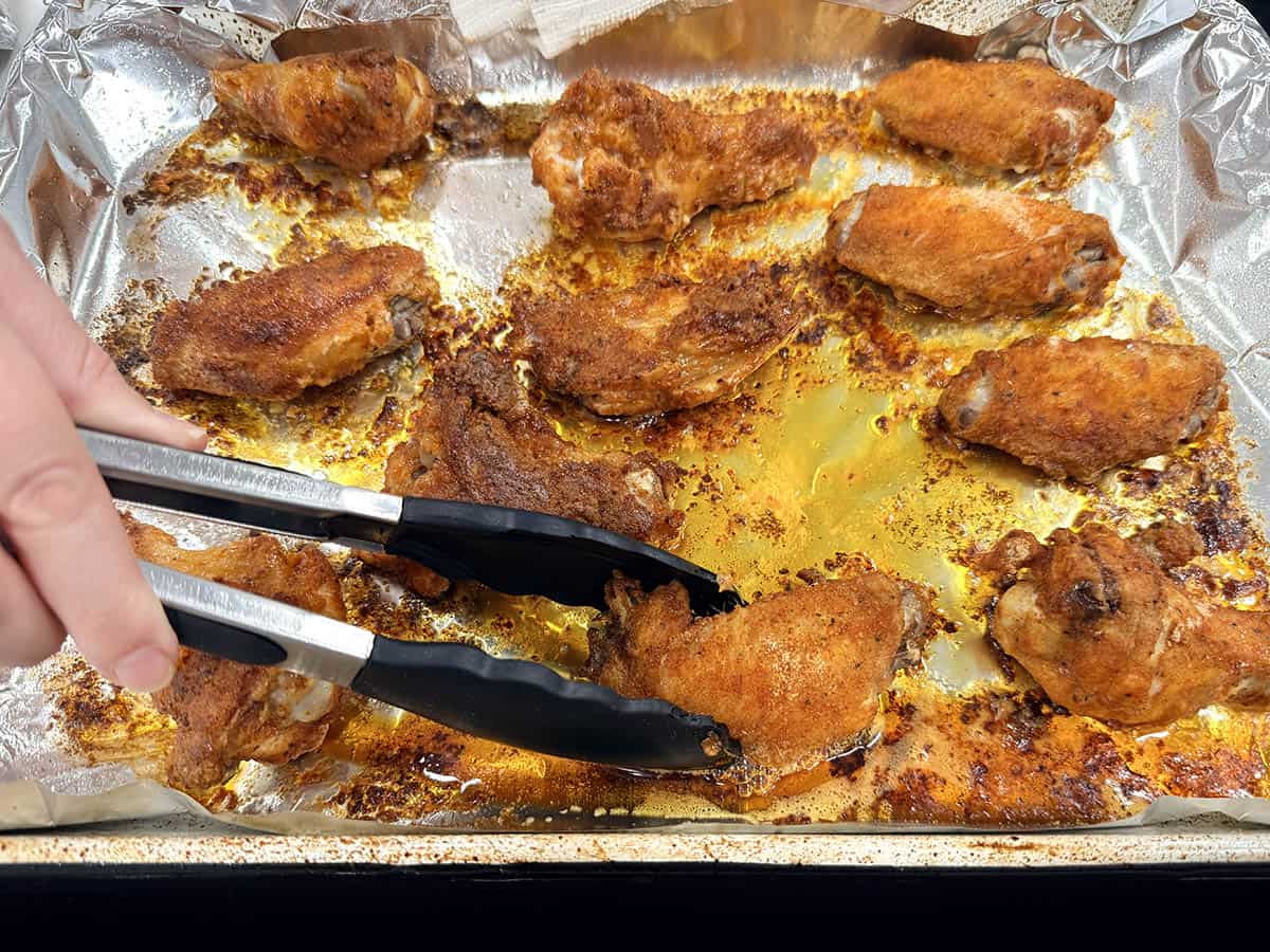 Tongs flipping a crispy baked chicken wing on the baking sheet halfway through cooking.