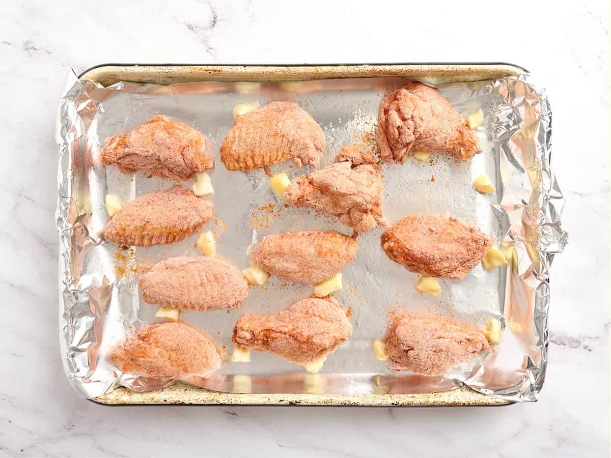 Flour coated chicken wings arranged on a foil lined baking sheet with butter pieces between them, ready to bake.