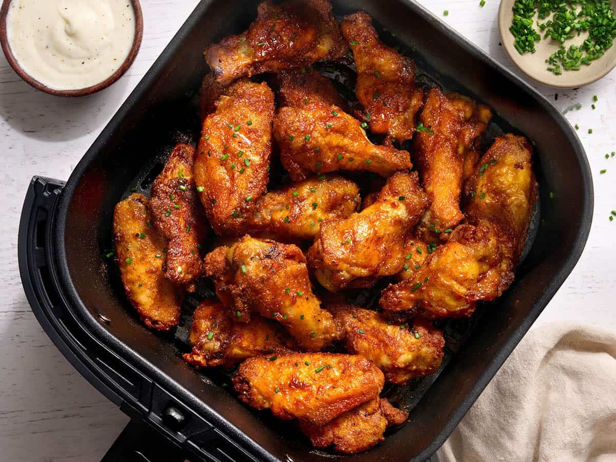 Overhead view of crispy air fryer chicken wings garnished with chopped chives in an air fryer basket, served with a bowl of ranch dressing and a small dish of chives