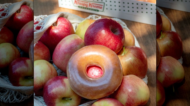 An apple cider donut surrounded by apples with a Krispy Kreme box in the background.
