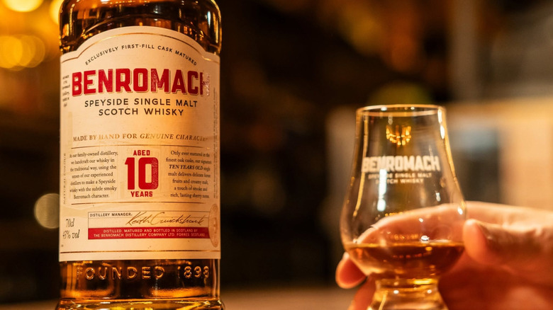 A close up of a bottle of Benromach 10-year next to a person reaching out for a filled glasss