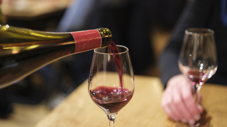 Red wine being poured into a glass while someone holds a mostly empty glass out of focus in the background.