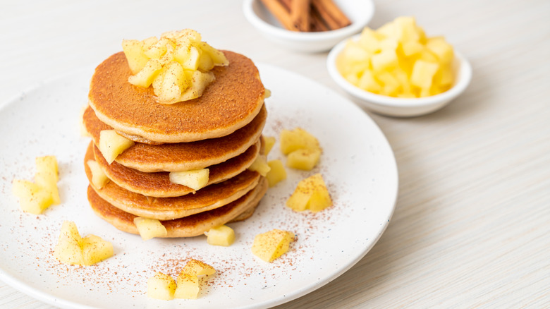White plate with stack of mini pancakes topped with diced apples and cinnamon with small white bowls of diced apples and cinnamon sticks in background.