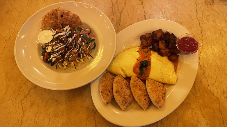 A Cheesecake Factory Mediterranean Bowl and Shakshuka Omelette sit in dishes next to each other on a marble table.