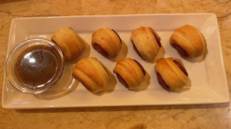 Seven pigs in a blanket bites sit on a white tray on a marble table.