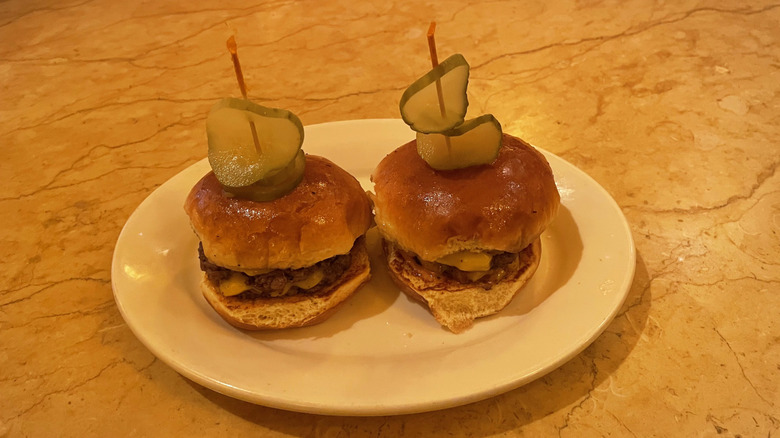 Two small smashburgers with pickle slices on a spear sit on a white dish on top of a marble table.