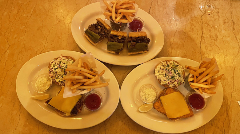 Three sandwiches (two fish, one cheesesteak) sit on white dishes on a marble tabletop.