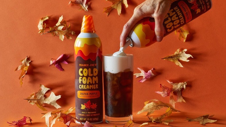 A hand dispensing Trader Joe's Salted Maple Cold Foam Creamer onto iced coffee with a background of fall leaves