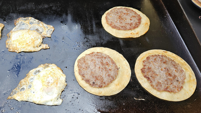 Two fried eggs and three tortillas on a griddle with sausage smashed into them and cooked