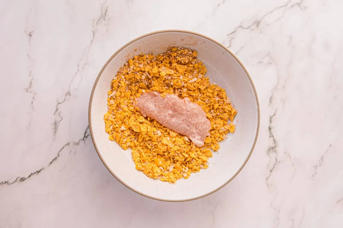 Raw chicken tenderloin resting on the cornflake mixture before being pressed and coated
