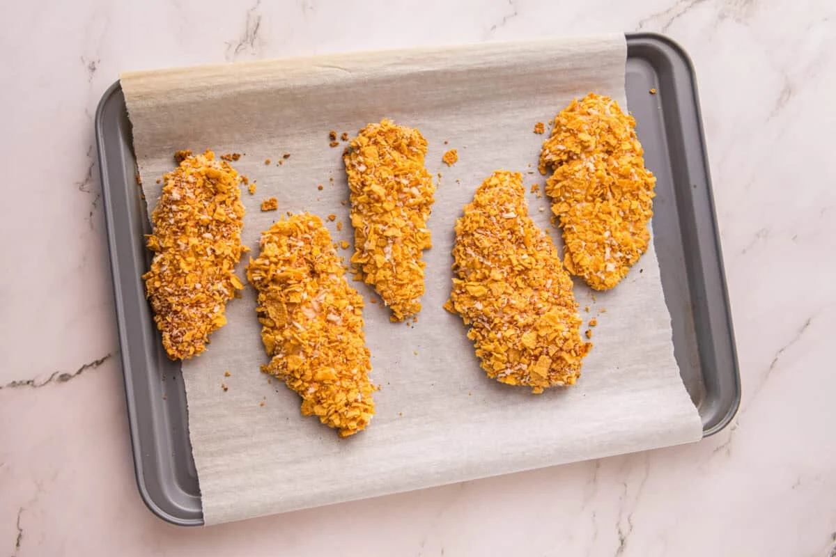 Five cornflake-coated chicken tenders arranged on a parchment-lined baking sheet before baking