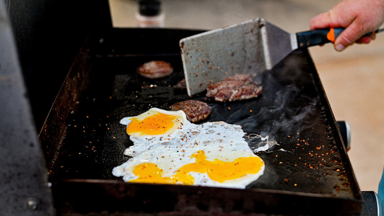 A person holding a large spatula cooking seasoned eggs and sausage patties on a flat top grill.