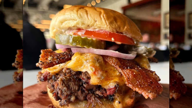 Massive brisket sandwich topped with melty cheese, bacon, tomato, pickle, and red onion on a soft bun
