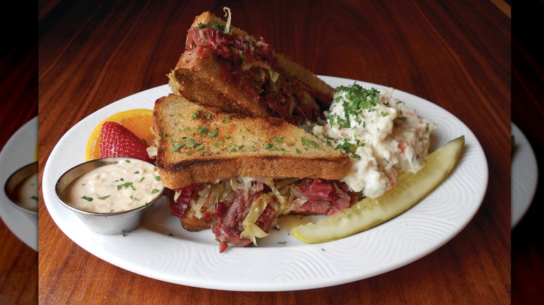 Reuben sandwich on toasted bread with corned beef and sauerkraut spilling out, on a white plate with a side of aioli, pickle, fruit, and potato salad