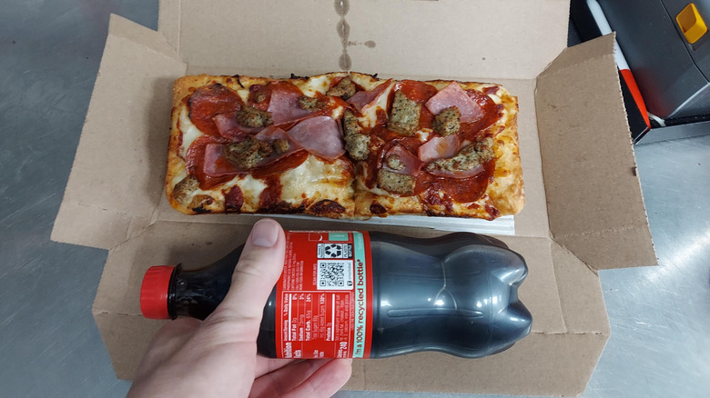 A Domino's Domio topped with ham pepperoni and sausage sitting in a box, its size being compared to a bottle of Coca-Cola held by a hand