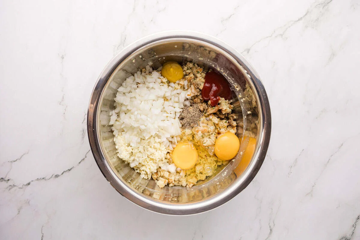 Diced onion, garlic, eggs, ketchup, and seasonings added to softened stuffing mix in a bowl