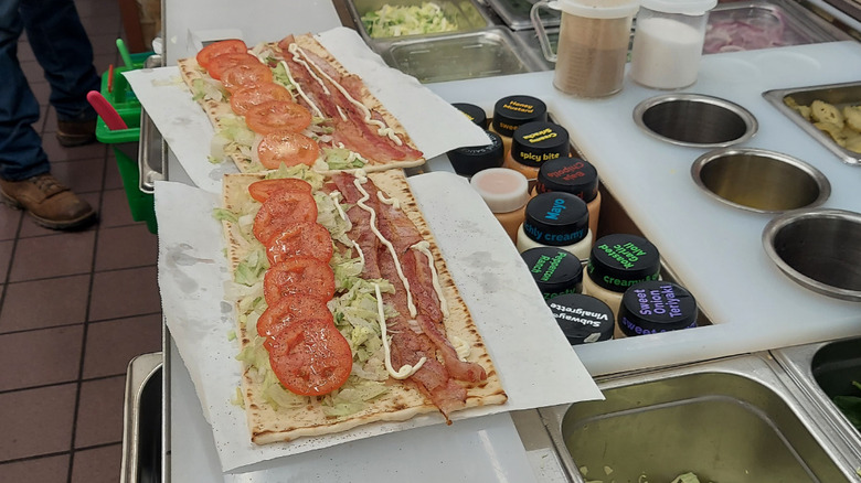 Two BLT flatbreads being made at a prep station in a Subway next to assorted sauces