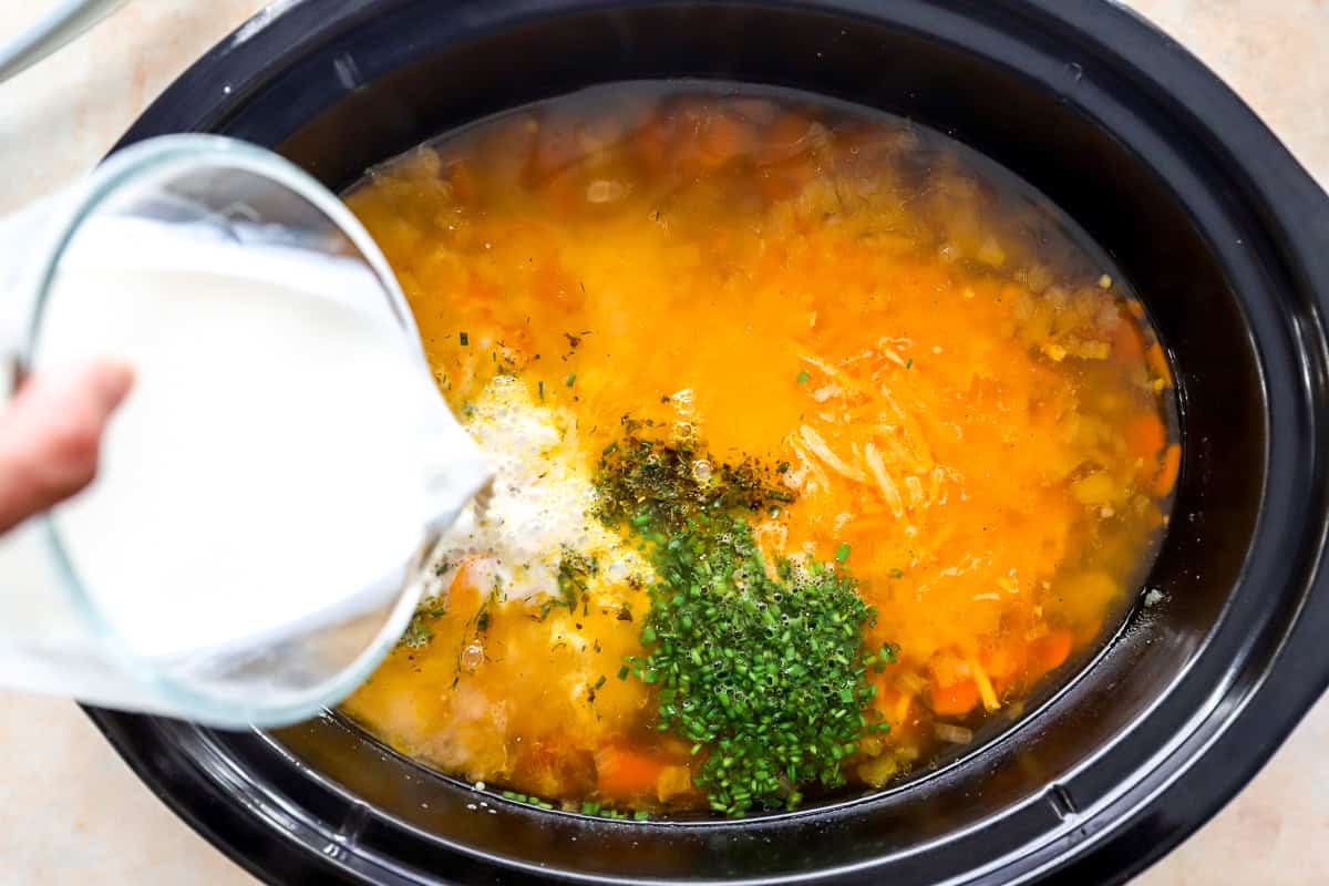Fat-free half-and-half being poured into a slow cooker with shredded cheddar, chopped chives, and dried herbs on top of the potato soup.