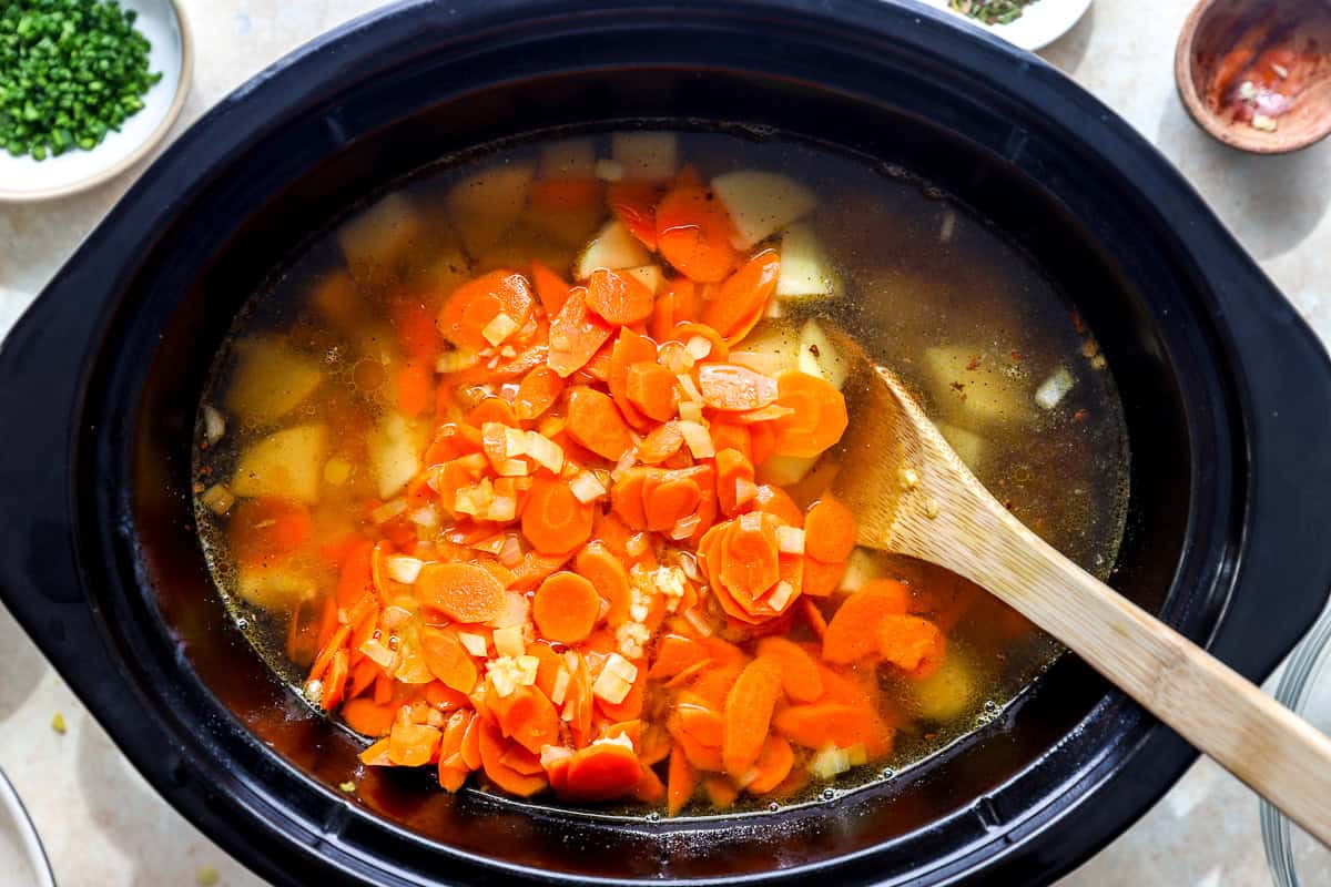 Sautéed carrots, onion, and garlic added to cubed potatoes and chicken broth in a slow cooker, with a wooden spoon.