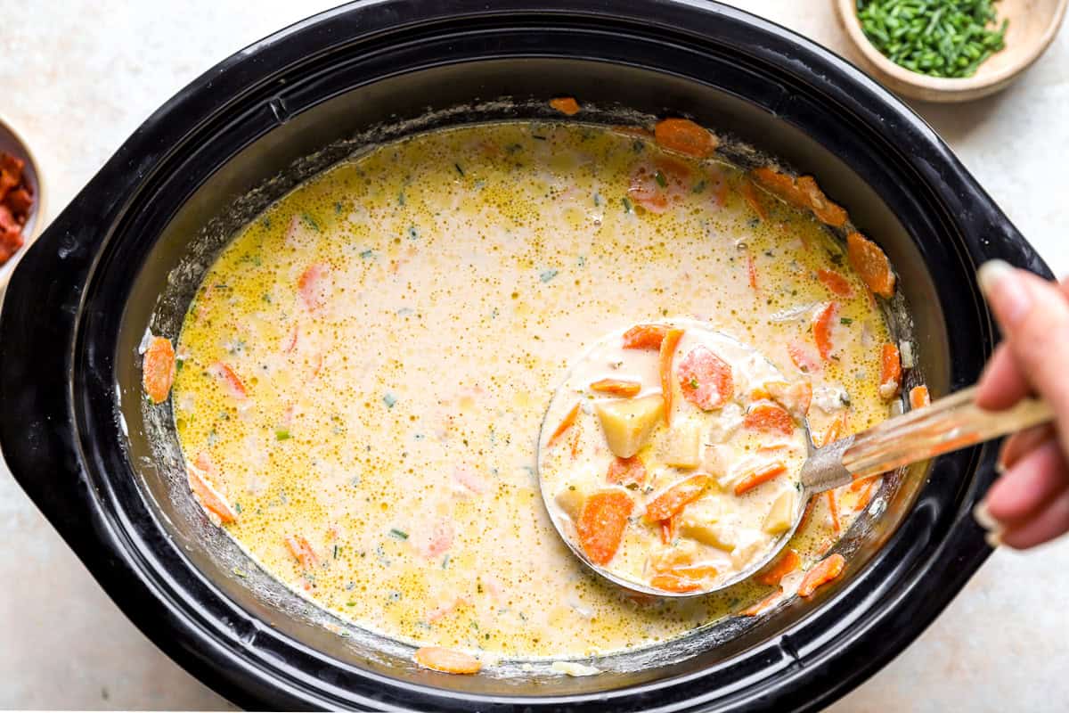 A ladle lifting creamy potato soup with chunks of potato and carrot from a slow cooker.