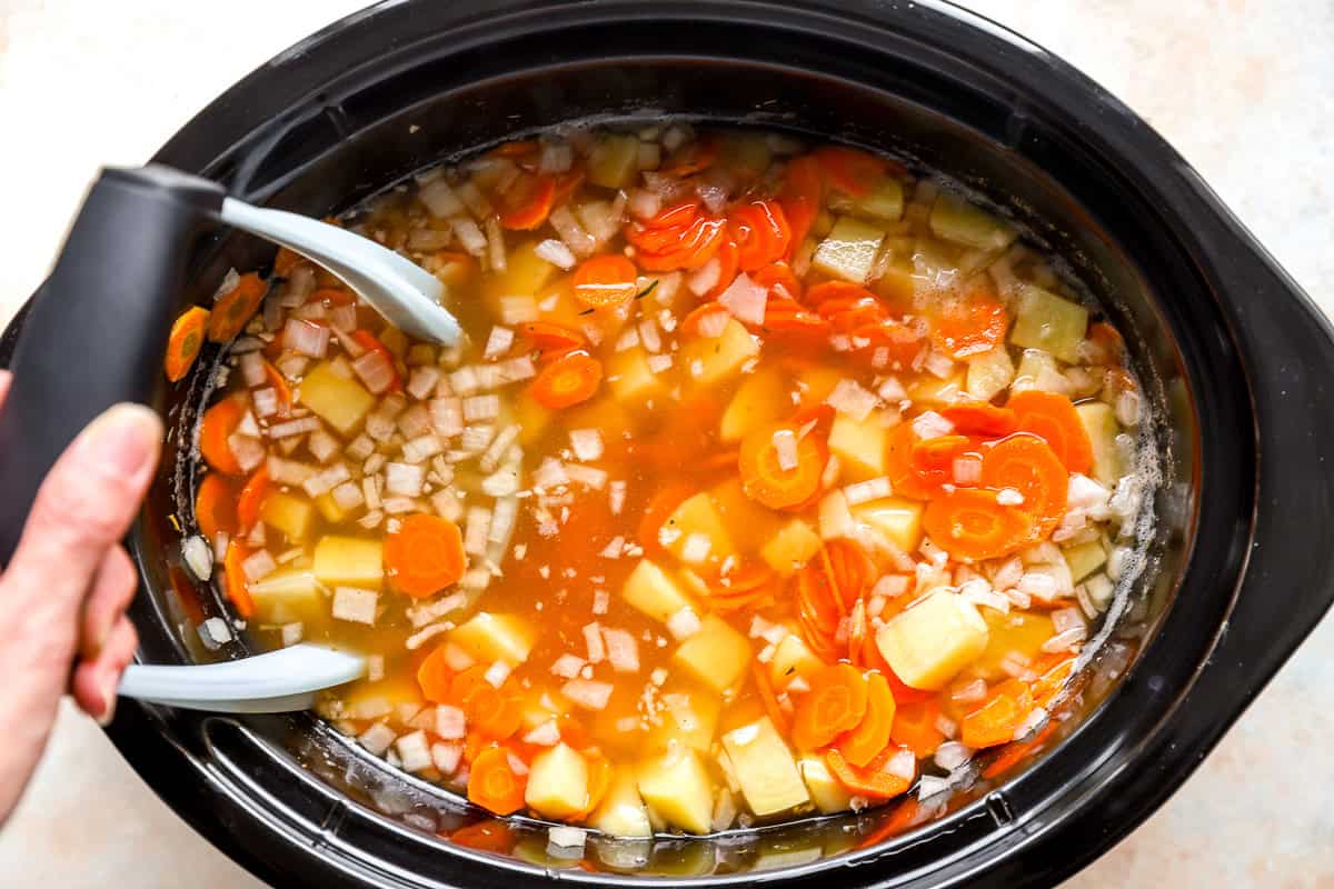 A potato masher pressing into cooked potatoes, carrots, and onion in broth inside a slow cooker.