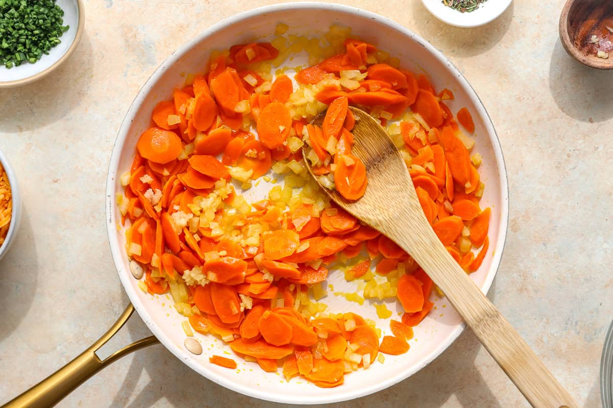 Sliced carrots, diced onion, and minced garlic sautéing in a white skillet with a wooden spoon.