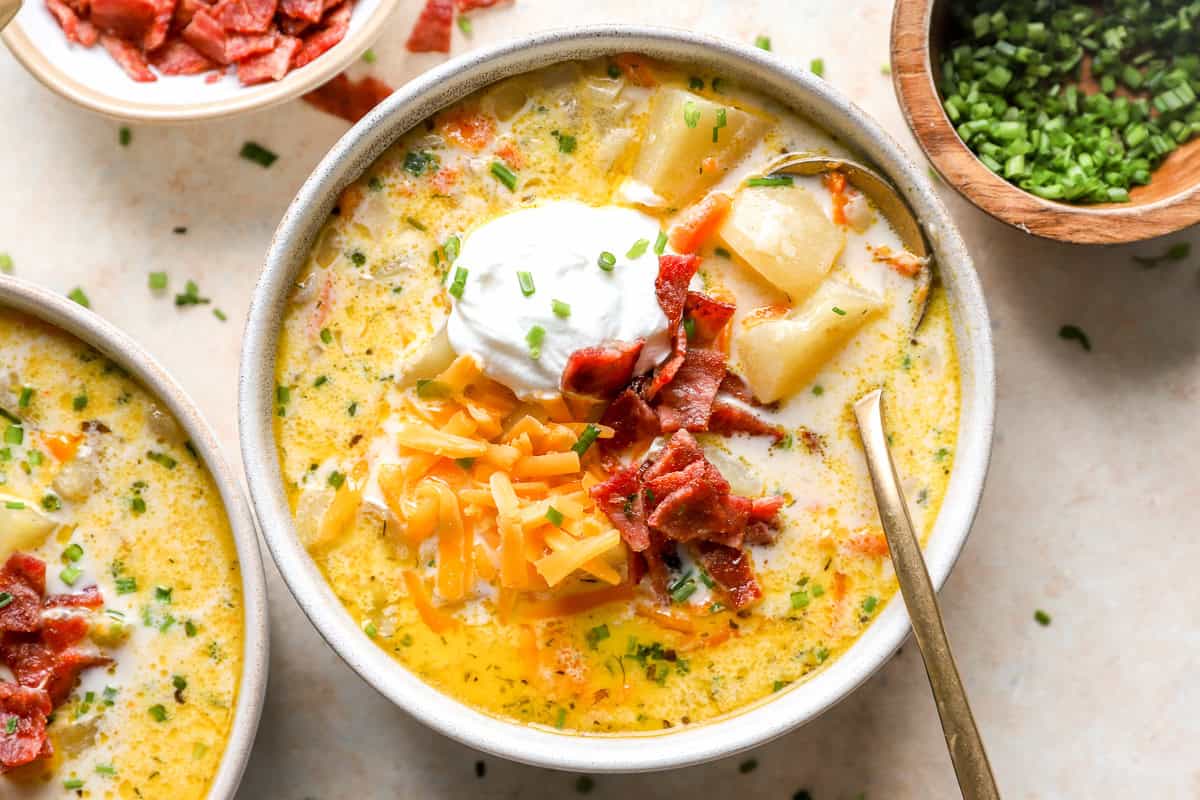 A bowl of crockpot potato soup topped with sour cream, turkey bacon, shredded cheddar, and chives, with a small bowl of chives on the side.