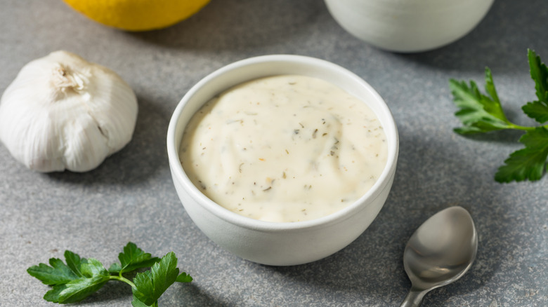 A bowl of ranch dressing in white ramekin next to head of garlic, herbs, and spoon