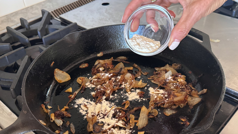 Hand pouring small bowl of flour over caramelized onions in skillet